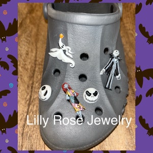 Op de afbeelding: Een grijze schoen met verschillende Nightmare Before Christmas-thema schoenbedels. De bedels omvatten Jack Skellington, Sally, Zero de spookhond en een gezicht van Jack Skellington. De schoenbedels zijn bevestigd aan een grijze schoen met gaten. De tekst "Lilly Rose Jewelry" is op de schoen gedrukt.