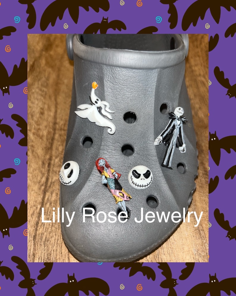 Op de afbeelding: Een grijze schoen met verschillende Nightmare Before Christmas-thema schoenbedels. De bedels omvatten Jack Skellington, Sally, Zero de spookhond en een gezicht van Jack Skellington. De schoenbedels zijn bevestigd aan een grijze schoen met gaten. De tekst "Lilly Rose Jewelry" is op de schoen gedrukt.