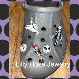 Op de afbeelding: Grijze schoen met verschillende bedels van personages uit Nightmare Before Christmas. De bedels zijn Jack Skellington, Sally, Zero de spookhond en Oogie Boogie. De schoen heeft de tekst "Lilly Rose Jewelry" op de onderkant.