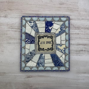 May include: A square mosaic tile with a blue and white pattern. The tile has a white border with a blue and white pattern. The center of the tile has a white square with the word "SHINE" in black lettering.
