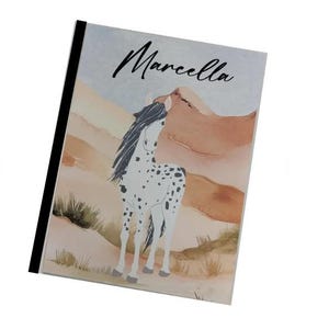 May include: Notebook with a watercolor illustration of a white horse with black spots, set against a backdrop of tan and brown mountains. The name "Marcella" is written in cursive at the top. The notebook has a black spine.