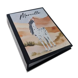 May include: A black binder with a watercolor design of a horse in a desert landscape. The binder has the name "Marcella" printed on the front.