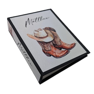 May include: A black binder with a white cover featuring a watercolor illustration of cowboy boots and a cowboy hat. The name "Matthew" is written in cursive on the cover.