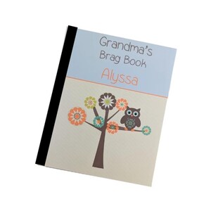 May include: A personalized notebook titled "Grandma's Brag Book" with the name "Alyssa" in orange. The cover features a tree with colorful flower designs and a brown owl perched on a branch. The notebook has a black spine.