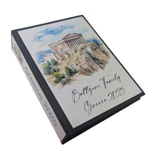 May include: A personalized photo album with a watercolor illustration of a Greek temple on the cover. The album is black with the text "Battzeri Family Greece 2025" in elegant script.