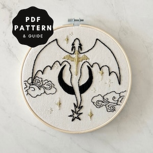 May include: Embroidered hoop art featuring a black and gold dragon flying through a crescent moon with clouds and stars. The hoop is made of wood and has a white fabric background.