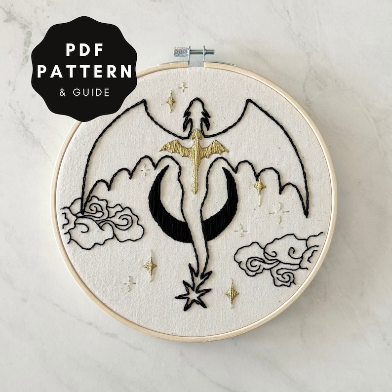 May include: Embroidered hoop art featuring a black and gold dragon flying through a crescent moon with clouds and stars. The hoop is made of wood and has a white fabric background.