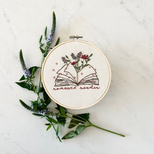 May include: Embroidered hoop art featuring an open book with flowers and the text "romance reader".