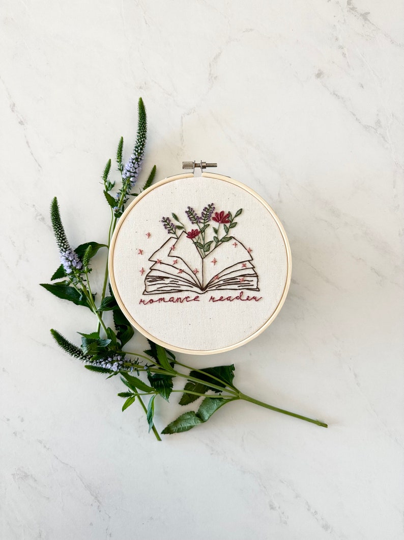 May include: Embroidered hoop art featuring an open book with flowers and the text "romance reader".