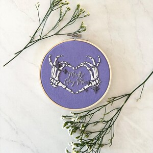 May include: Purple embroidery hoop with a heart formed by two skeleton hands. The text "Mortally Gay Men" is embroidered inside the heart.
