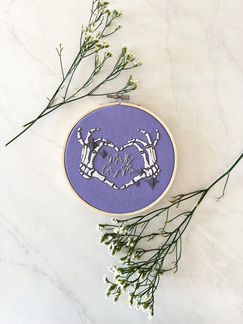 May include: Purple embroidery hoop with a heart formed by two skeleton hands. The text "Mortally Gay Men" is embroidered inside the heart.