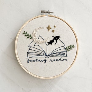 May include: Embroidered hoop art featuring an open book with a crescent moon and stars above it. The book is surrounded by green leaves and the text "fantasy reader" is stitched below.