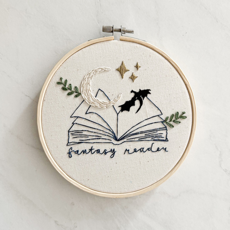 May include: Embroidered hoop art featuring an open book with a crescent moon and stars above it. The book is surrounded by green leaves and the text "fantasy reader" is stitched below.