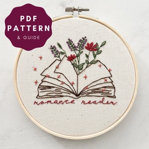 May include: Embroidered hoop art featuring an open book with flowers growing from it. The book is surrounded by small stars and the text "romance reader" is stitched below the book.