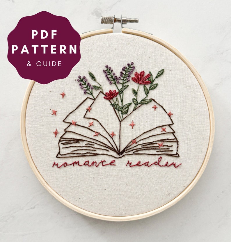 May include: Embroidered hoop art featuring an open book with flowers growing from it. The book is surrounded by small stars and the text "romance reader" is stitched below the book.