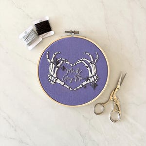 May include: Purple embroidery hoop with a design of two skeleton hands forming a heart shape. The text "Mostly Gray Morn" is embroidered inside the heart.