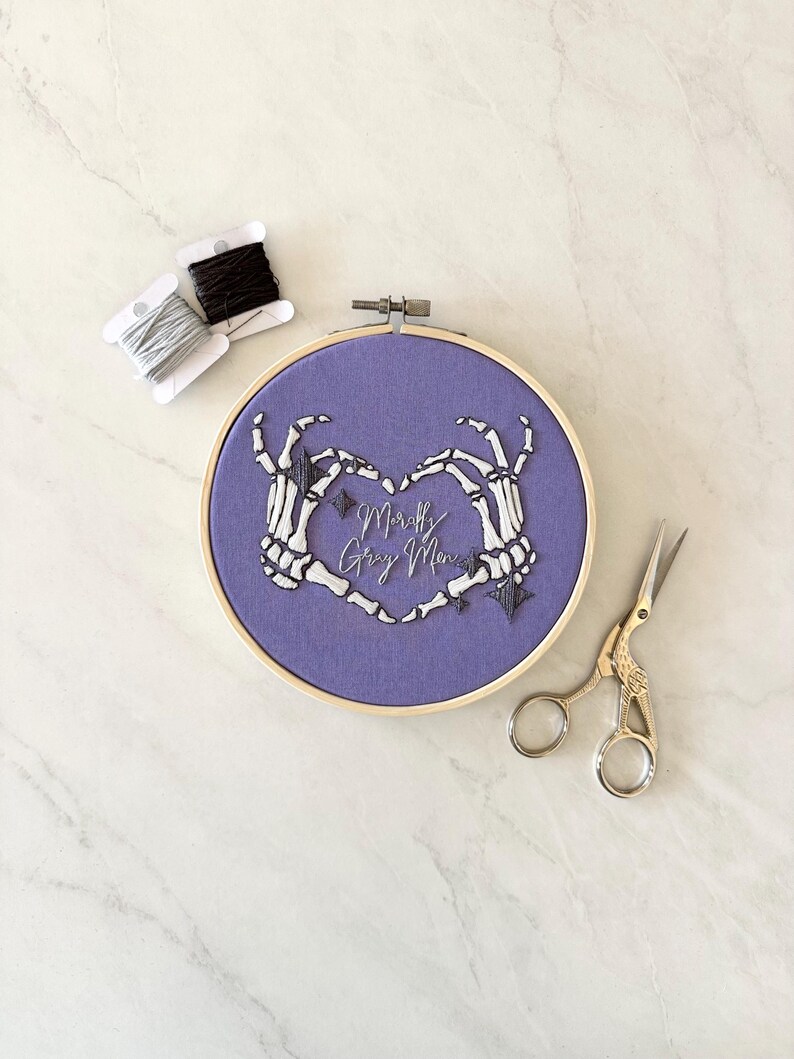 May include: Purple embroidery hoop with a design of two skeleton hands forming a heart shape. The text "Mostly Gray Morn" is embroidered inside the heart.