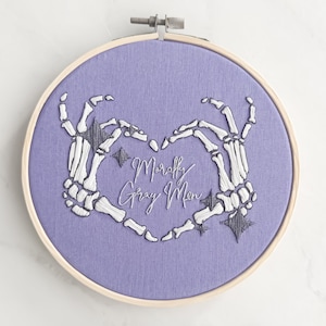 May include: Embroidered hoop art with a purple background featuring two white skeleton hands forming a heart shape. The text "Morally Gray Men" is embroidered in white inside the heart.