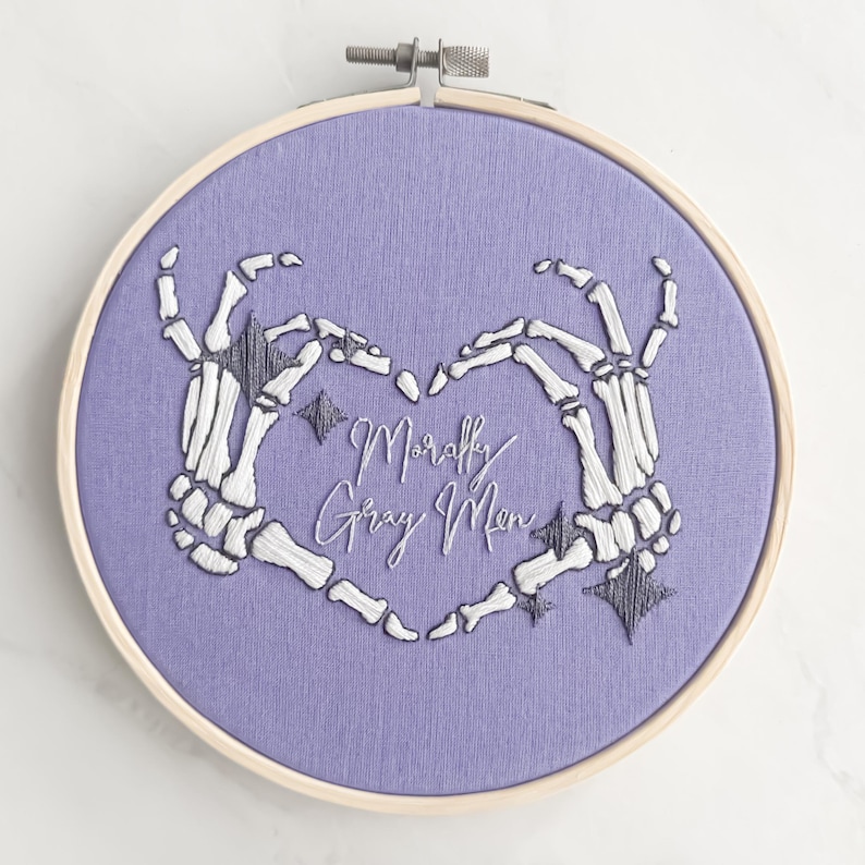 May include: Embroidered hoop art with a purple background featuring two white skeleton hands forming a heart shape. The text "Morally Gray Men" is embroidered in white inside the heart.
