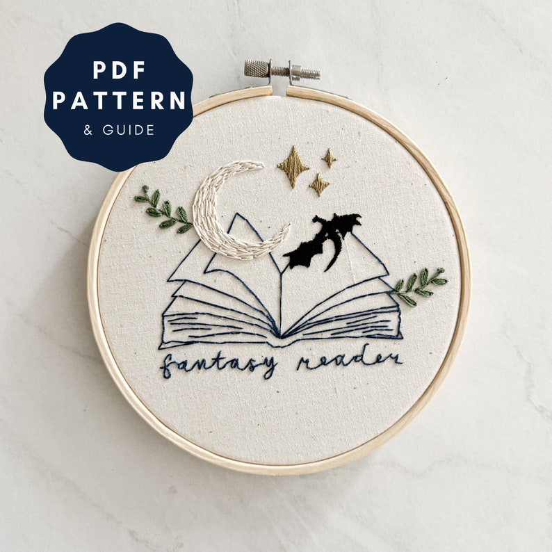 May include: Embroidered hoop art featuring a book with a crescent moon and stars above it. The book is open and the text "fantasy reader" is stitched below it.