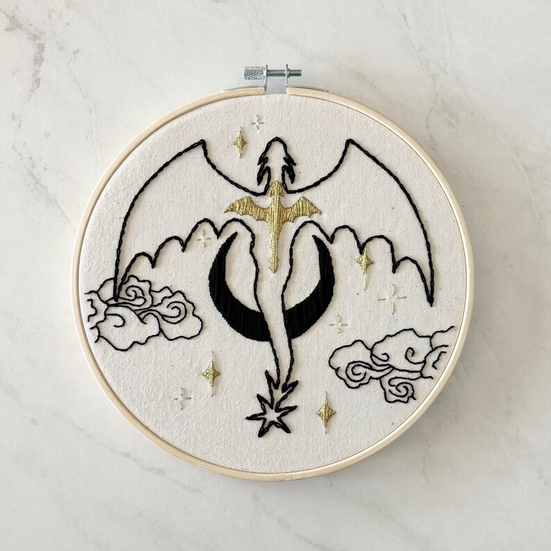 May include: Embroidered hoop art featuring a black and gold dragon flying against a white background with black clouds and gold stars.