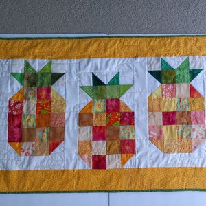 Può includere: Un quilt patchwork giallo e verde con tre motivi a forma di ananas. Gli ananas sono realizzati con diversi quadrati di tessuto colorati.
