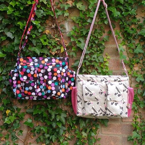 Può includere: Due borse a tracolla colorate appese a un gancio. La borsa a sinistra ha uno sfondo nero con pois colorati e più tasche. La borsa a destra ha uno sfondo bianco con un motivo di pattini a rotelle in bianco e nero e una tasca a rete rosa.