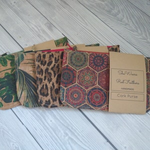 May include: Four cork purses with different patterns. The purses are lined up in a row on a wooden surface. The patterns are: tropical leaves, leopard print, geometric pattern, and a geometric pattern with a tag that reads "She Wears Red Feathers Handmade Cork Purse".