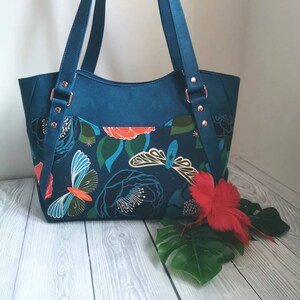 May include: A teal blue tote bag with a floral print featuring butterflies and flowers. The bag has two straps and a pocket on the front.