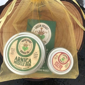 Puede incluir: Dos pequeñas latas redondas de bálsamo herbal natural en una bolsa de organza amarilla. La lata más grande está etiquetada como "Arnica Muscle Rub" y la lata más pequeña está etiquetada como "Everything Salve". Ambas latas tienen un logotipo de corazón verde con una corona y el texto "Green Heart Herbal Co."