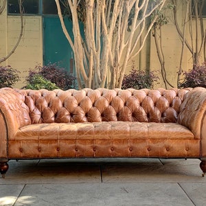 May include: A brown leather tufted sofa with rolled arms and wooden legs. The sofa is in a rustic setting with trees and a yellow wall in the background.