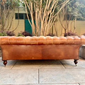 May include: A brown leather sofa with tufted back and rolled arms. The sofa has wooden legs and is sitting on a stone patio.