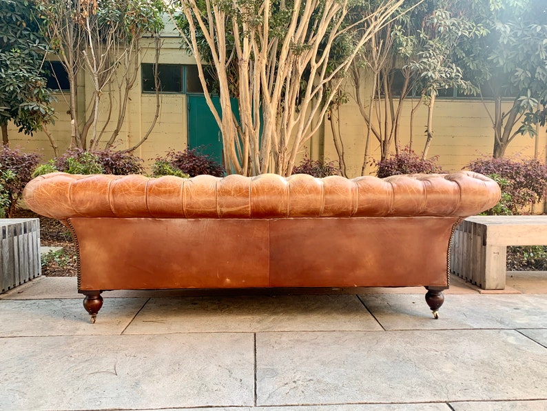 May include: A brown leather sofa with tufted back and rolled arms. The sofa has wooden legs and is sitting on a stone patio.