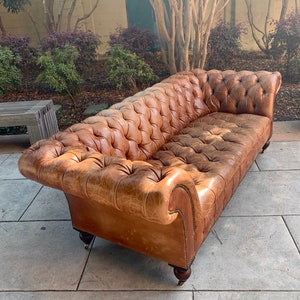 May include: A brown leather tufted sofa with a distressed finish. The sofa has a classic design with rolled arms and button tufting. The sofa is sitting on a concrete patio.