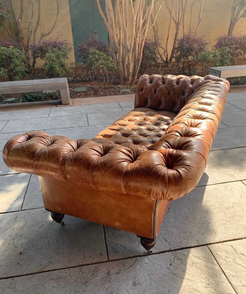 May include: A brown leather tufted sofa with a distressed finish. The sofa has a rolled back and arms and sits on four wooden legs with casters.