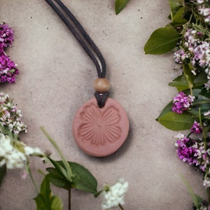May include: A pink ceramic pendant with a floral design hangs from a brown cord. The pendant is surrounded by white and purple flowers.