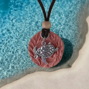 May include: A pink ceramic pendant with a silver turtle charm hanging from a brown leather cord. The pendant has a textured surface and the turtle charm is detailed with raised lines.