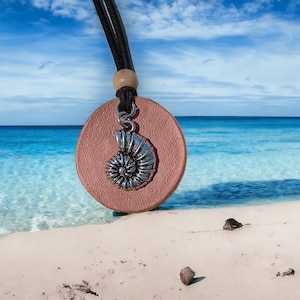 May include: A pink leather pendant necklace with a silver seashell charm. The necklace is photographed on a white sandy beach with blue ocean water in the background.