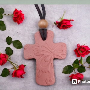 May include: A terracotta cross necklace with a butterfly design. The cross is hanging on a white background with red roses around it.
