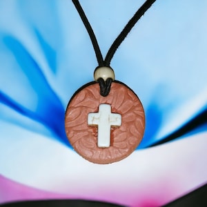 May include: A brown ceramic pendant with a white cross design hangs from a black cord. The pendant is on a blue and pink background.