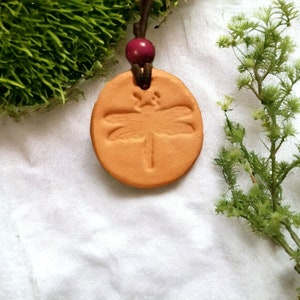 May include: A terracotta pendant necklace with a dragonfly design. The necklace has a brown cord and a burgundy bead.