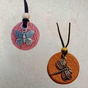 May include: Two necklaces with round pendants. The pendant on the left is pink with a silver butterfly charm. The pendant on the right is orange with a gold dragonfly charm.