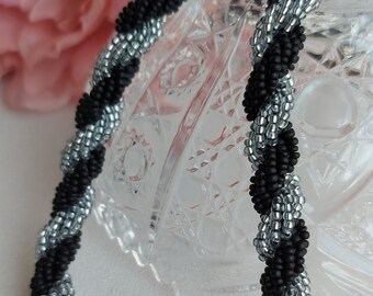 May include: A black and silver beaded necklace with a twisted design. The necklace is made of small, round beads that are woven together in a spiral pattern.