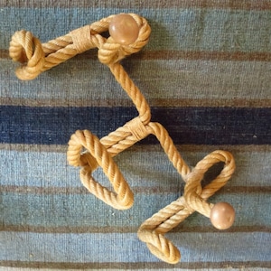 May include: A rope coat rack with three wooden knobs. The rack is made of natural brown rope and is displayed on a striped rug.