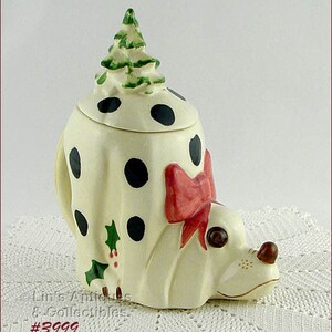 May include: A ceramic Dalmatian dog cookie jar with black spots, a red bow, and a green Christmas tree on top. The jar has a cream-colored base and is decorated with holly and berries.