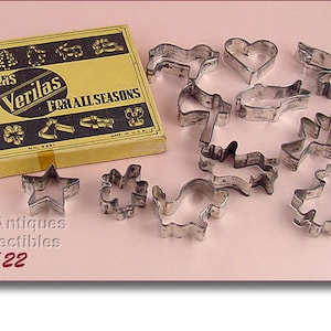 May include: A vintage set of metal cookie cutters in a yellow box. The box says "Cooky Cutters Verilas For All Seasons". The cutters are in the shape of a star, heart, bird, fish, and other designs.