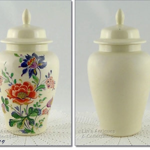 May include: Cream-colored ceramic jar with a lid. One side features a floral design with red, blue, and green accents. The other side is plain. The jar has a bulbous shape and a matching lid with a small finial.