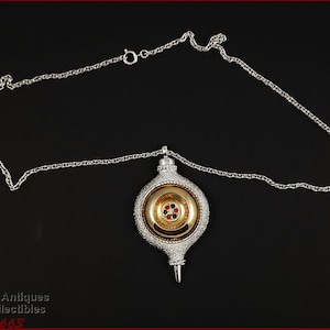 May include: Silver-toned necklace with a pendant. The pendant is an elongated, ornate shape with a gold-colored center and a silver-colored outer layer. The center has red and green accents. The necklace is on a black background.