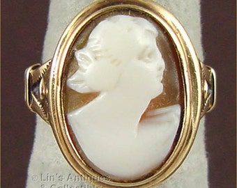 May include: A gold ring featuring an oval cameo with a carved profile of a woman. The cameo is set in a gold frame with decorative detailing on the sides. The ring is displayed on a white surface.