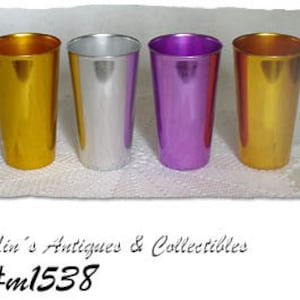 May include: Four metallic tumblers in gold, silver, and purple hues. The cylindrical cups are arranged side-by-side on a white, textured surface. The text "Lin's Antiques & Collectibles" and "#ml1538" are visible.